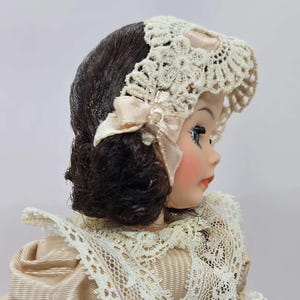 May include: A vintage doll with dark brown hair and a cream-colored lace bonnet. The doll has rosy cheeks, red lips, and is wearing a cream-colored dress with lace trim. A light pink bow adorns the bonnet.