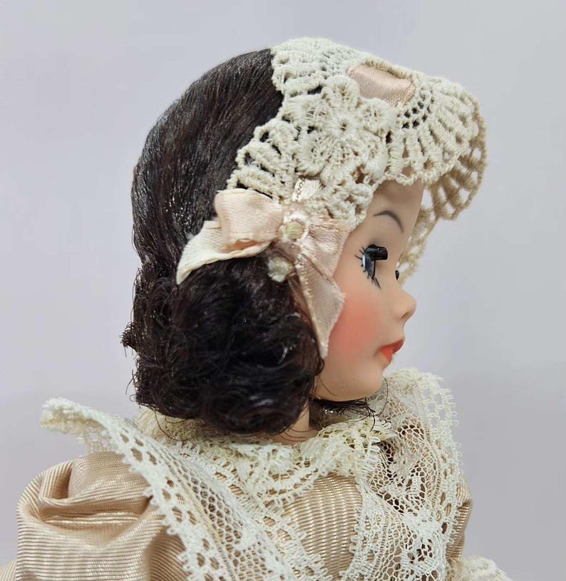 May include: A vintage doll with dark brown hair and a cream-colored lace bonnet. The doll has rosy cheeks, red lips, and is wearing a cream-colored dress with lace trim. A light pink bow adorns the bonnet.