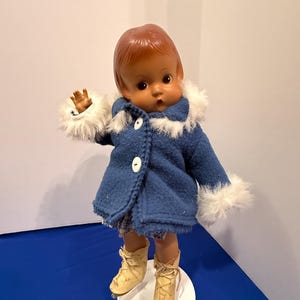 Op de afbeelding: Een vintage pop met bruin haar en ogen, gekleed in een blauwe jas met witte bontafwerking en witte knopen. De pop draagt ook een geruite korte broek en crèmekleurige schaatsen. De pop staat op een witte cirkelvormige basis.