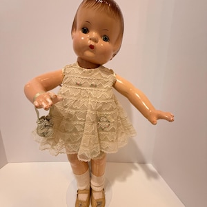 Könnte beinhalten: Vintage Puppe mit bemaltem Gesicht, trägt ein cremefarbenes Spitzenkleid und passende Schuhe. Die Puppe hält eine kleine Handtasche und hat kurze, braune Haare. Die Puppe steht auf einem transparenten Kunststoffständer.