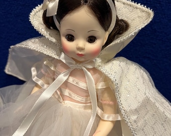 Può includere: Una bambola di porcellana vestita con un abito bianco con un corpetto rosa e un mantello bianco. La bambola ha i capelli castani, gli occhi marroni e le guance rosate. La bambola tiene un biglietto che dice "Biancaneve".