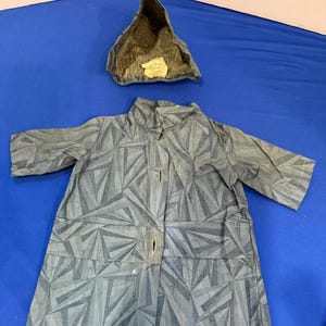 Könnte beinhalten: Ein grauer Mantel mit geometrischem Dreiecksmuster, ausgestattet mit Kragen, langen Ärmeln und einer Knopfleiste vorne. Eine passende spitze Kapuze mit einem beigen Innenetikett liegt darüber und zeigt den Text Elite Creations.
