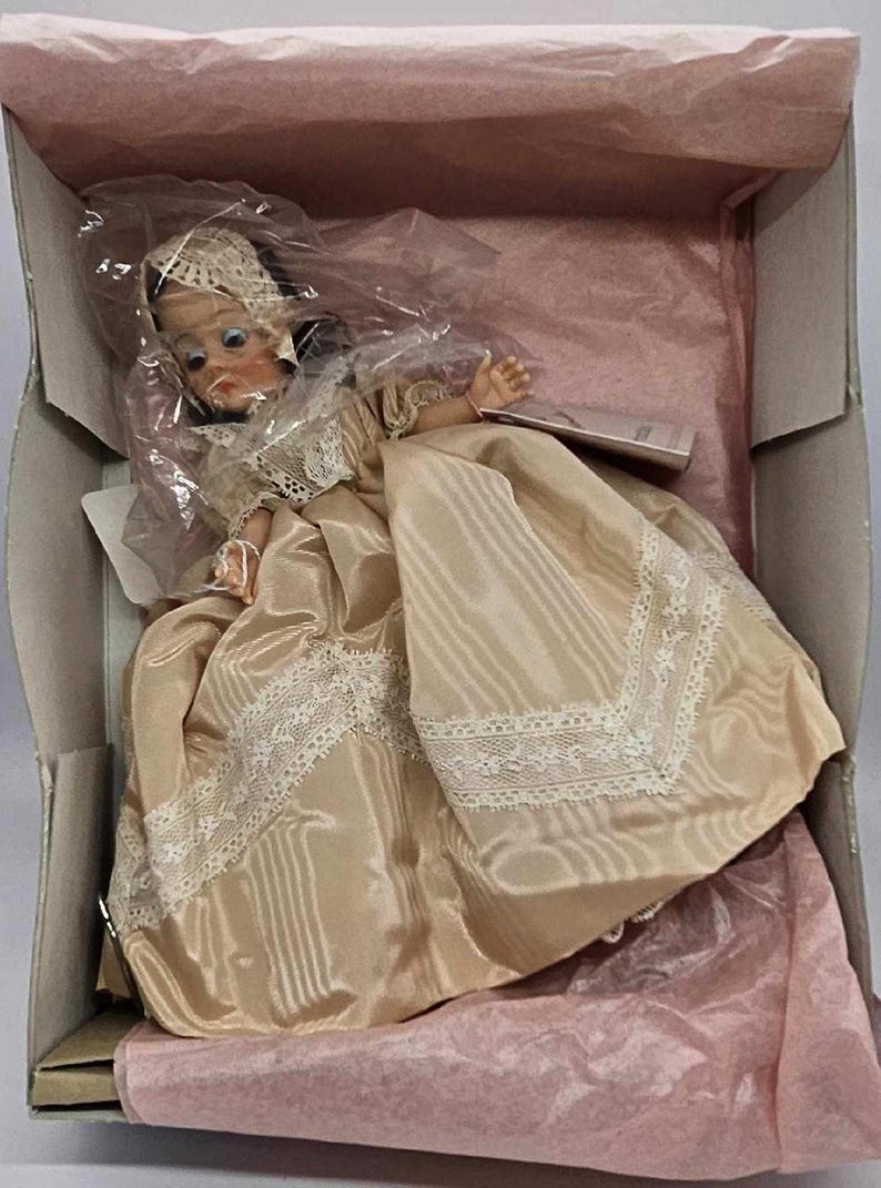 May include: A vintage doll in a box, dressed in a satin gown with lace trim. The doll has a bonnet and is packaged in a box with pink tissue paper.