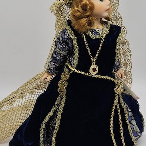 May include: A porcelain doll with auburn hair and a gold-colored crown and necklace. The doll wears a long, dark blue velvet dress with gold trim and a matching gold cape. The doll has a serene expression and is posed in a three-quarter view.