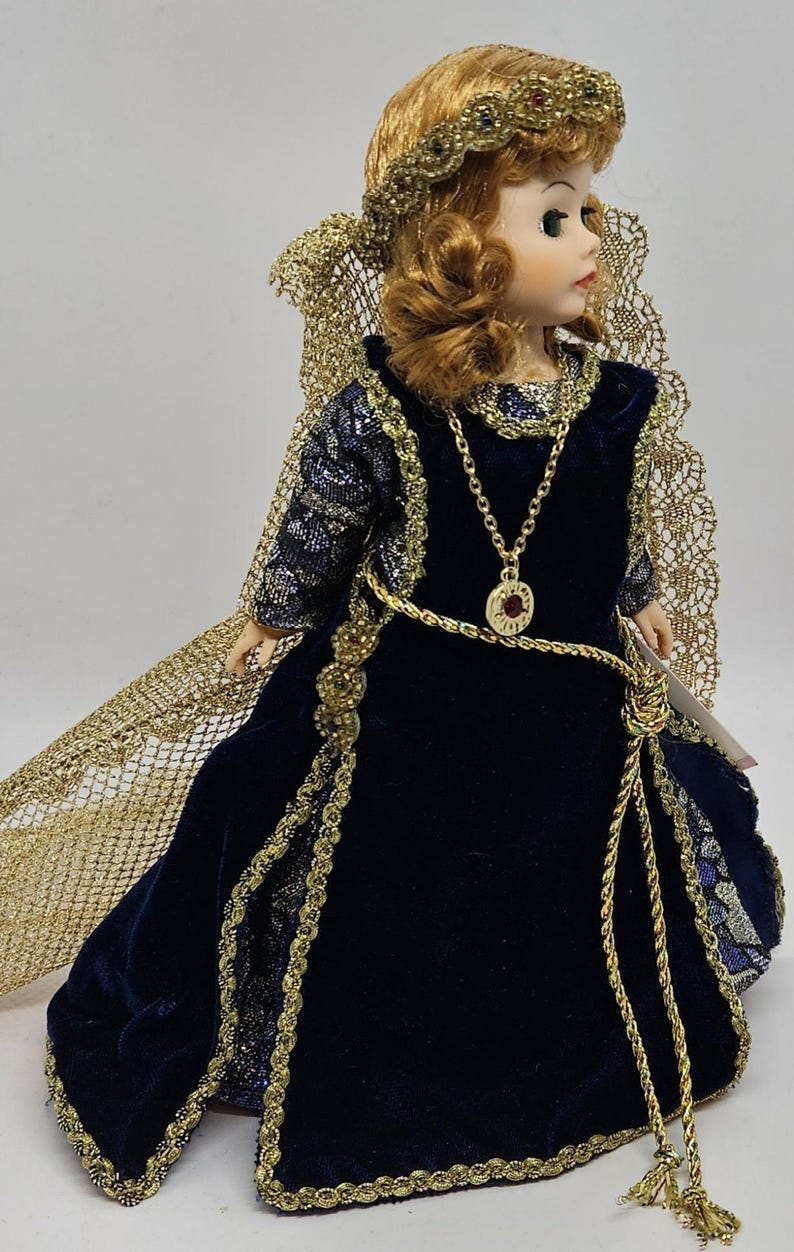 May include: A porcelain doll with auburn hair and a gold-colored crown and necklace. The doll wears a long, dark blue velvet dress with gold trim and a matching gold cape. The doll has a serene expression and is posed in a three-quarter view.