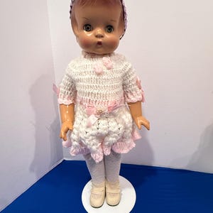 Puede incluir: Una muñeca vintage con una diadema rosa y un vestido de ganchillo blanco y rosa. La muñeca tiene una expresión dulce y lleva medias blancas y zapatos color crema. El vestido presenta lazos y detalles rosas, añadiendo un toque de encanto.