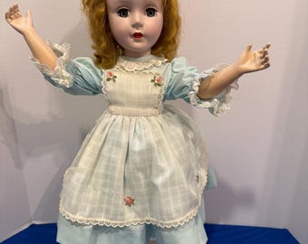 Può includere: Una bambola con capelli ricci castano-rossicci, occhi azzurri e labbra rosse. La bambola indossa un vestito azzurro con maniche bordate di pizzo bianco e un grembiule a quadri bianchi con piccoli ricami floreali rosa. Indossa scarpe nere.