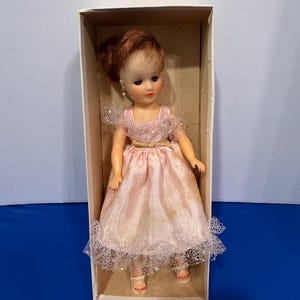 May include: A vintage doll in a cardboard box. The doll has red hair styled in an updo, wearing a pink dress with lace trim and a gold belt. The doll is wearing white sandals with red painted toenails. The doll has dangling earrings.