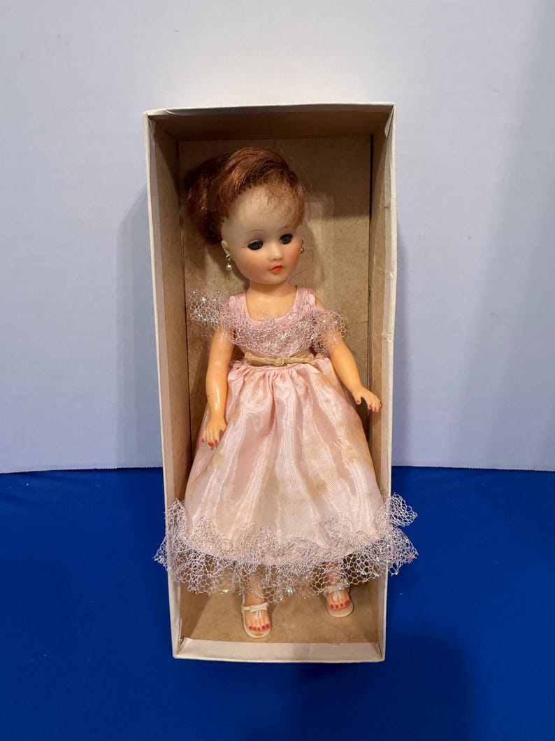 May include: A vintage doll in a cardboard box. The doll has red hair styled in an updo, wearing a pink dress with lace trim and a gold belt. The doll is wearing white sandals with red painted toenails. The doll has dangling earrings.