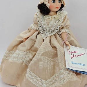 May include: A vintage Madame Alexander Portrettes doll. The doll wears a beige satin dress with white lace trim and a matching bonnet. The doll has dark hair, blue eyes, and a tag that reads "Madame Alexander Portrettes."