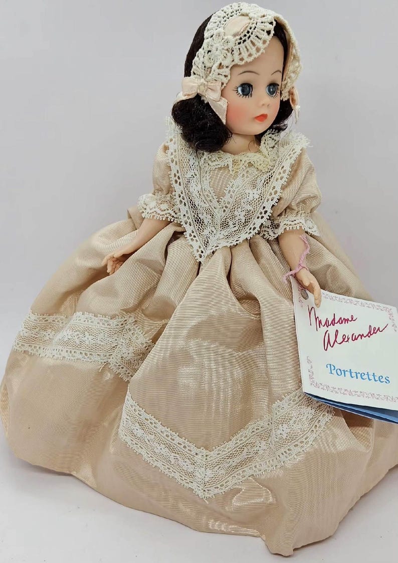 May include: A vintage Madame Alexander Portrettes doll. The doll wears a beige satin dress with white lace trim and a matching bonnet. The doll has dark hair, blue eyes, and a tag that reads "Madame Alexander Portrettes."