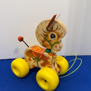 Könnte beinhalten: Vintage Fisher-Price "Fancy Pony" Spielzeug zum Ziehen. Das Spielzeugpferd hat einen hölzernen Kopf und Körper mit einem bemalten Design. Es hat gelbe Räder und eine grüne Schnur. Das Pony hat einen roten Sattel und eine rote Kugel an einer Metallstange.