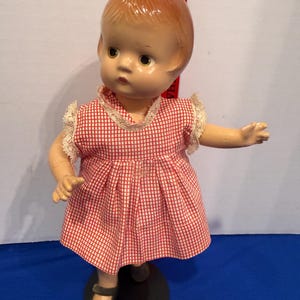 May include: A vintage doll with a painted face, wearing a red and white checkered dress with lace trim. The doll has a molded hairstyle and is posed on a black stand. The doll's shoes are black.