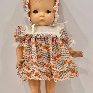 Può includere: Una bambola vintage con capelli modellati color marrone rossiccio, che indossa una cuffietta e un vestito abbinati con stampa floreale e bordo in pizzo bianco. La bambola ha un colletto bianco, calzini a coste color crema e scarpe bianche con lacci.