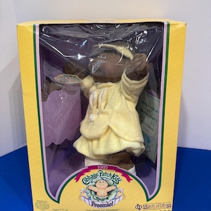 Op de afbeelding: Een 1985 Cabbage Patch Kids Preemie pop met een donkere huidskleur, gekleed in een geel jack met capuchon en een kleine konijntjesafbeelding op de voorkant. De pop zit in een gele en paarse kartonnen doos met doorzichtig plastic, voorzien van de tekst Cabbage Patch Kids Preemie en March of Dimes.