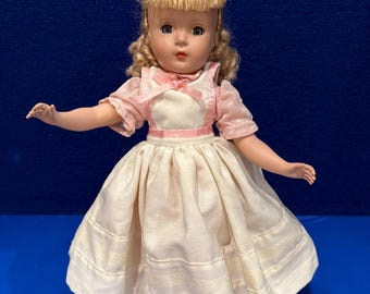 Pode incluir: Uma boneca vintage com cabelo loiro, olhos azuis e um vestido rosa e branco. A boneca está usando um vestido branco com um corpete rosa e uma saia de bolinhas rosa e brancas.
