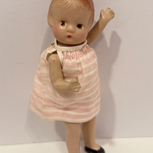Può includere: Bambola vintage con viso dipinto, labbra rosse e capelli corti castano-rossicci. La bambola indossa un vestito a righe rosa e bianco e scarpe nere. Un braccio è alzato. Un giocattolo classico.