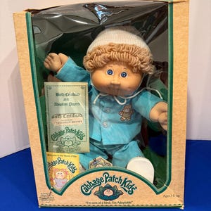 Peut inclure: Une poupée Cabbage Patch Kids au teint hâlé, aux yeux bleus et aux cheveux en laine brun clair, portant un bonnet en tricot blanc et un pyjama bleu clair à manches longues avec un motif d'ours. La poupée est dans une boîte avec son certificat.