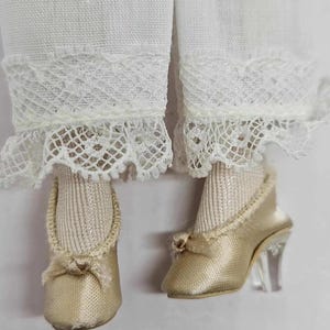 May include: Close-up of doll clothing featuring white lace-trimmed pants and gold satin shoes. The shoes have clear heels and small bows. The doll's socks are a light beige color.