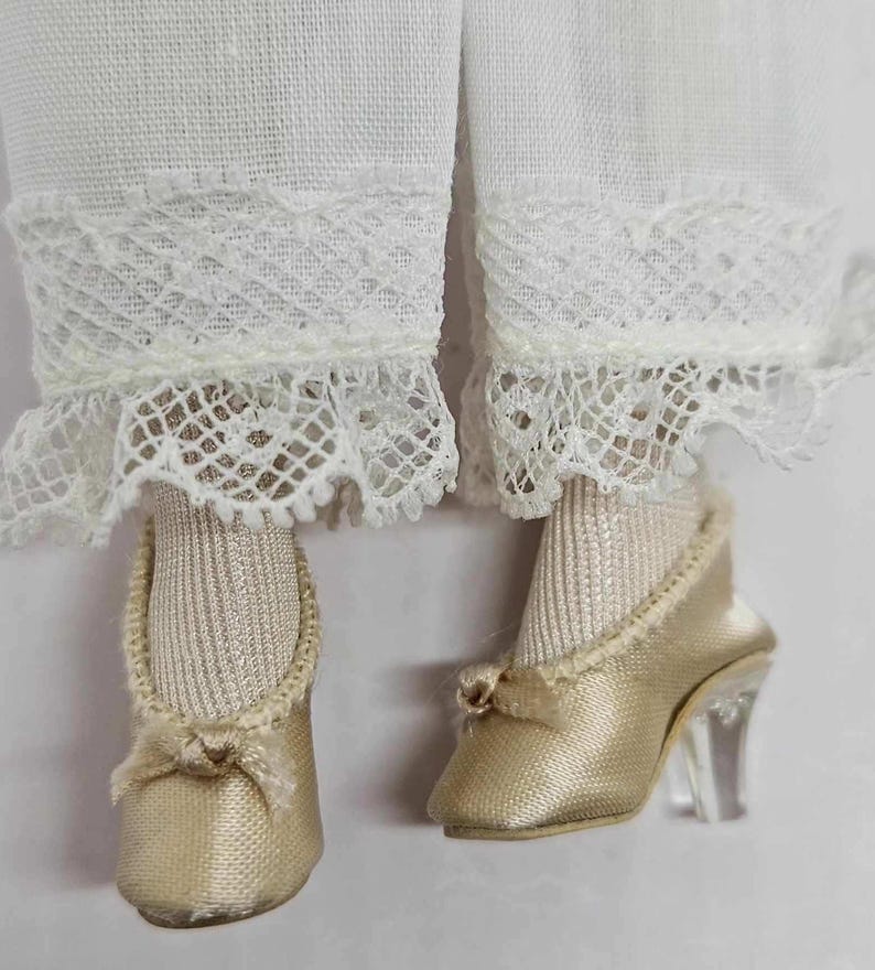 May include: Close-up of doll clothing featuring white lace-trimmed pants and gold satin shoes. The shoes have clear heels and small bows. The doll's socks are a light beige color.