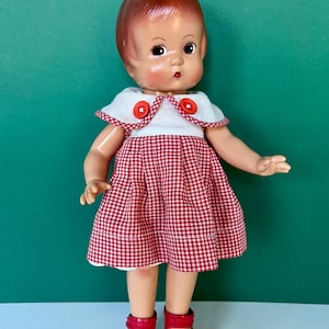 Peut inclure: Une poupée aux cheveux brun-roux et aux traits peints, vêtue d'un col blanc avec deux boutons rouges et d'une robe à carreaux vichy rouges et blancs. La poupée porte des chaussures rouges avec de petites roues fixées aux semelles, comme des patins.