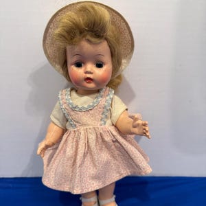 May include: A vintage doll with blonde hair, wearing a pink dress with a white pattern and a matching hat. The doll has painted facial features, including rosy cheeks and red lips. The doll is wearing white shoes.
