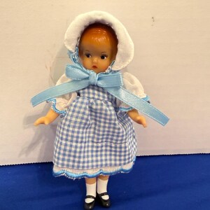 May include: A small doll wearing a blue and white checkered dress, white bonnet, and black shoes. The doll has a blue ribbon tied in a bow at the neck. The doll's dress has a white trim.