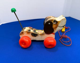 Könnte beinhalten: Ein klassisches Fisher-Price Little Snoopy Nachziehspielzeug in Form eines weiß-braun gefleckten Hundes mit vier leuchtend roten Kunststoffrädern. Das Spielzeug hat einen langen Federschwanz mit grüner Kugelspitze und eine rote Zugschnur.