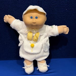 Può includere: Una bambola Cabbage Patch Kid vestita con un completo bianco in spugna con dettagli gialli. La bambola ha gli occhi azzurri e un cappello bianco.