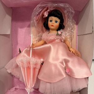 May include: A vintage doll in a pink satin dress, matching hat, and a lace-trimmed parasol. The doll has black hair and is packaged in a pink box. The dress features a tulle underskirt and a small rose accent.