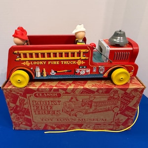 Könnte beinhalten: Ein rotes Vintage-Spielzeug "Looky Fire Truck" mit gelben Rädern, einer silbernen Glocke und zwei Spielzeug-Feuerwehrleuten. Das Spielzeug steht auf einem Karton mit dem Text "Looky Fire Truck" und "Toy Town Museum".