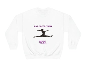 May include: White sweatshirt with a black silhouette of a gymnast in a split pose. The text "EAT, SLEEP, TRAIN" is above the gymnast and the text "REPEAT" is below the gymnast. The text is in purple.