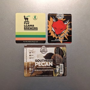 May include: Three beer coasters with different designs. The first coaster is green, yellow, and orange with a llama and the text "Fly Llama Brewing" and "Fly-PA India Pale Ale". The second coaster is black, red, and yellow with a fox and the text "Lazy Magnolia" and "Surf-X IPA". The third coaster is brown with a pecan and the text "Lazy Magnolia" and "Southern Pecan".