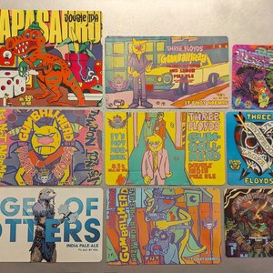 May include: A collection of nine colorful beer bottle labels featuring various designs, including a dinosaur, a skull, and a cartoon character. The labels are all rectangular and have a variety of colors and fonts.