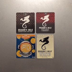 May include: Four different beer bottle labels featuring a dragon design. The labels are for Dragon's Milk Bourbon Barrel-Aged Stout, Dragon's Milk Solera, Tangerine Space Machine New England-Style India Pale Ale, and Dragon's Milk White Bourbon Barrel-Aged White Stout.