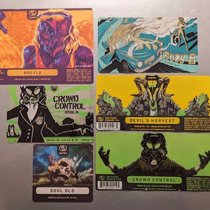 May include: Six colorful beer can labels with different illustrations and text. The labels feature a bear, a car, a skull, and a person wearing a gas mask. The text on the labels includes "Bro Flo", "Crowd Control", "Devil's Harvest", "Soul Glo", and "Suzy B".