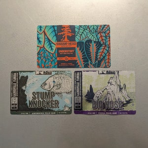 May include: Three beer bottle labels with different designs. The top label is blue and green with the text "Swamp Head Brewery" and "Understory" in white. The middle label is blue and green with a fish and the text "Stump Knocker" in white. The bottom label is green and purple with a tree and the text "Big Nose" in white.