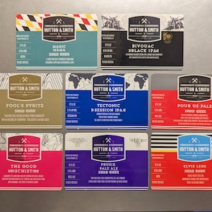 May include: Nine different beer labels from Hutton & Smith Brewing Company. The labels are all different colors and feature different designs. The beer names are: Magic Mama, Bivouac Black IPA, Fool's Pyrite, Tectonic Session IPA, Pour Us Pale, The Good Schist, Prusie Pale Ale, Elvis Leg.