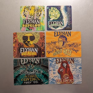 May include: Six square Elysian beer coasters with colorful graphics. Designs include a tiger, a cartoon artichoke, a man with sunglasses, a man with a brain graphic, a figure in water, and a figure in a red robe.