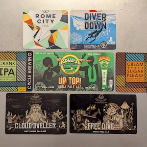 May include: A collection of nine colorful beer bottle labels featuring various designs and text, including "Crank IPA", "Rome City IPA", "Diver Down", "Dub's Brewing Company Up Top! India Pale Ale", "Cream ++++ Sugar Please", "Cloud Dweller Hazy India Pale Ale", and "Free Dive India Pale Ale".