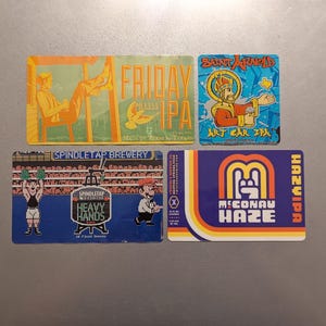 May include: Four colorful square stickers with different designs and text. The top left sticker is yellow and orange with a cartoon person sitting on a hammock. The text reads "Friday Miles IPA". The top right sticker is blue and yellow with a cartoon person wearing a crown and holding a beer mug. The text reads "Saint Arnold Art Car IPA". The bottom left sticker is blue and white with a cartoon person holding a weight. The text reads "Spindletap Brewery Heavy Hands IPA". The bottom right sticker is blue, yellow, and orange with a stylized "M" and the text "McConau Haze IPA".