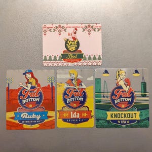 May include: Four colorful square coasters with illustrations of women and the text "Fat Bottom" in a circle. The coasters feature the names "Ruby", "Ida", "Knockout", and "Noel" with the corresponding beer styles.