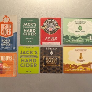 May include: Nine different hard cider labels with various flavors and brand names. The labels are colorful and feature illustrations of fruit, such as apples, pineapples, and mangoes.