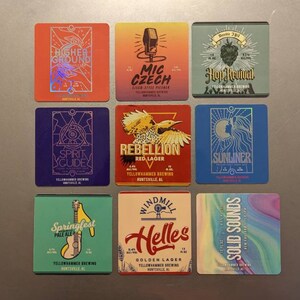 May include: A collection of nine square beer coasters with colorful designs and text. Each coaster features a unique graphic and the name of a beer, such as "Higher Ground," "Mic Czech," and "Hop Revival." The coasters are from Yellowhammer Brewing.