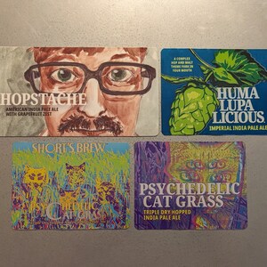May include: Four colorful beer coasters with different designs. The first coaster features a man with a mustache and glasses and the text "Hopstache American India Pale Ale with Grapefruit Zest". The second coaster features a green hop and the text "Huma Lupa Licious Imperial India Pale Ale". The third coaster features a psychedelic cat and the text "Short's Brew Psychedelic Cat Grass". The fourth coaster features a psychedelic cat and the text "Psychedelic Cat Grass Triple Dry Hopped India Pale Ale".