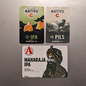 May include: Three beer labels with different designs. The first label is green and white with the text "COLORADO NATIVE" and "IPA". The second label is beige and white with the text "COLORADO NATIVE" and "PILS". The third label is white with a drawing of a man wearing a turban and the text "MAHARAJA IPA".