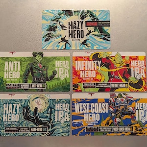 May include: Six different beer can labels featuring colorful illustrations of superheroes and the text "Hero IPA". The labels are arranged in two rows of three. The top row features "Anti Hero", "Infinity Hero", and "Hazy Hero". The bottom row features "West Coast Hero", "Hazy Hero", and "Hero IPA".