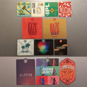 May include: A collection of colorful beer coasters with various designs and text. The coasters feature graphics like a gavel, a lantern, and abstract patterns. Text includes "Judge Juicy," "Light Lager," and "Camellia Blush Cider."