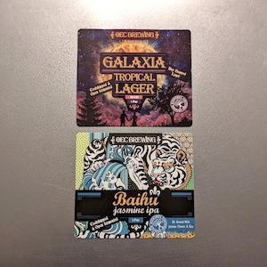 May include: Two beer coasters. The top coaster features the text "GALAXIA TROPICAL LAGER" with a starry background. The bottom coaster has the text "Baihu jasmine ipa" with a tiger illustration and blue wave design.