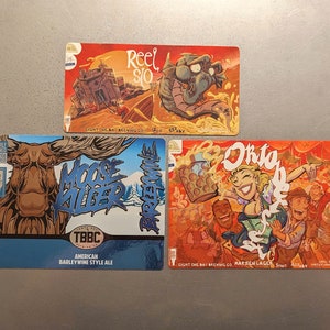 May include: Three colorful beer coasters with cartoon illustrations. The first coaster features a red background with a cartoon illustration of a building and a creature in the water. The second coaster features a blue background with a cartoon illustration of a moose and the text "Moose Killer". The third coaster features a red background with a cartoon illustration of a group of people and the text "Oktoberfest".